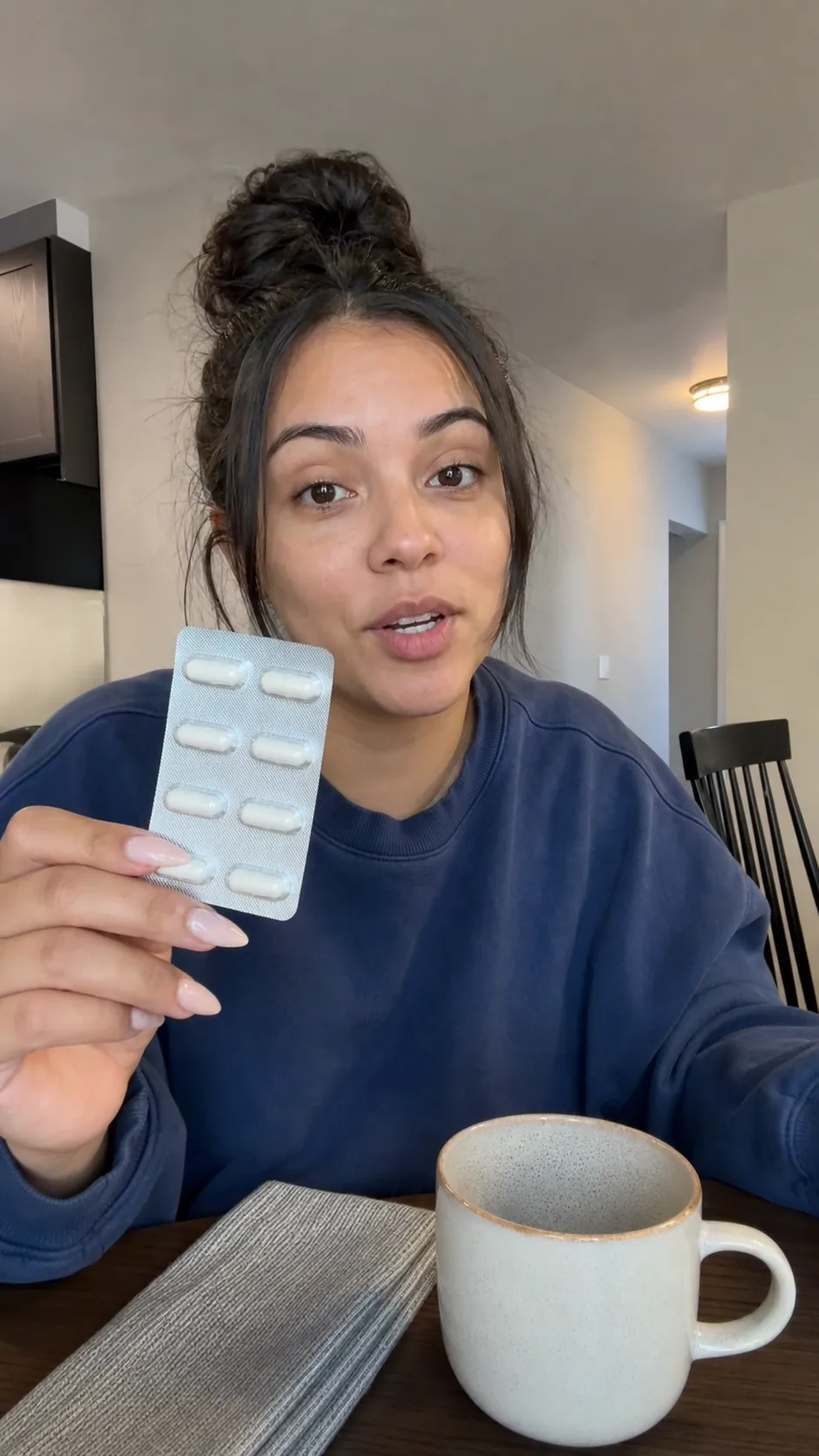 Woman at a dining table holds a plain capsule blister strip toward the camera.